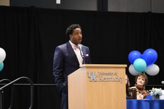a photograph of Dr. Swannie Jett speaking at a podium labeled “University of Kentucky”, on a stage of panelists