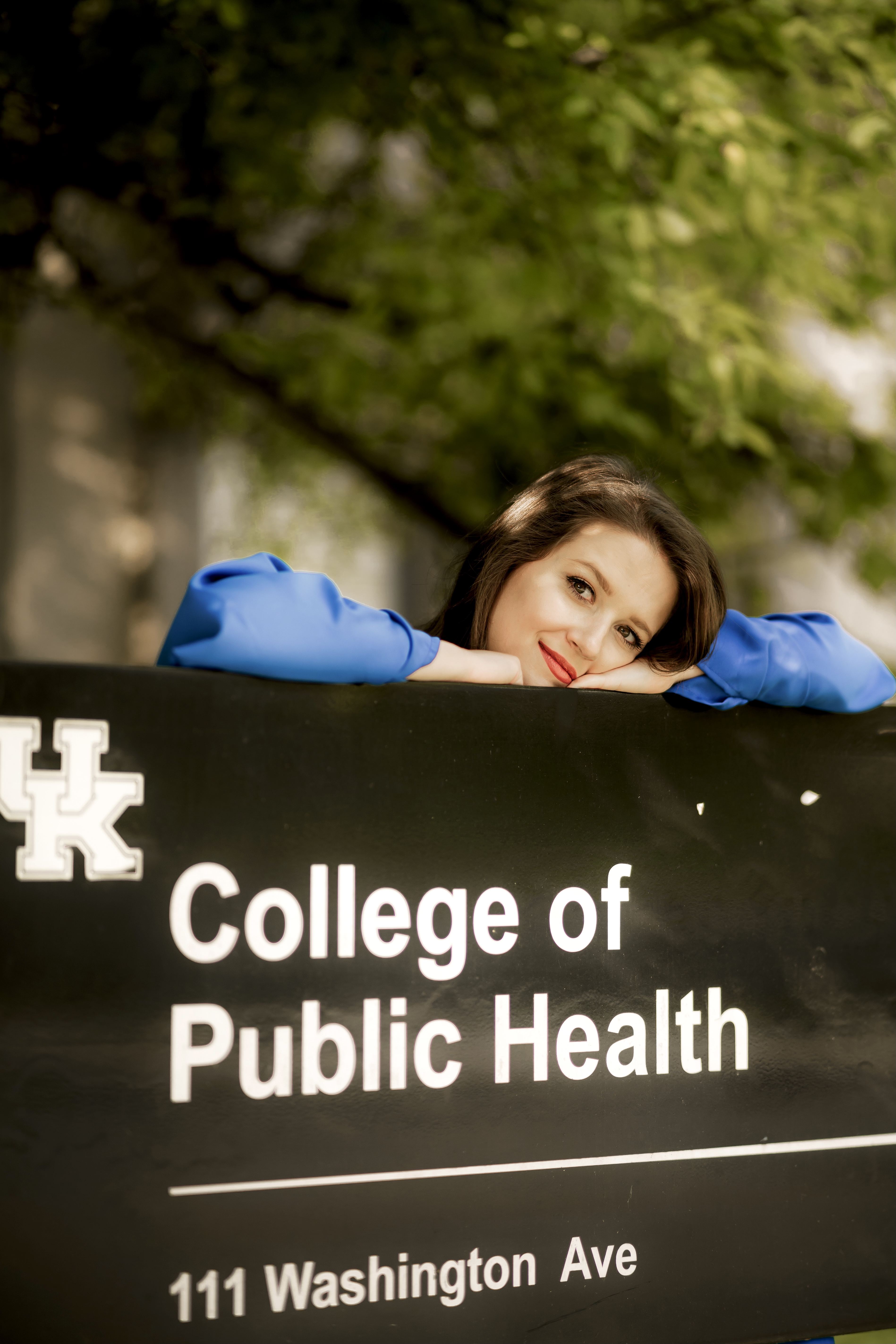 a photograph of Sarah Jane Robbins resting their head on a sign for the College of Public Health