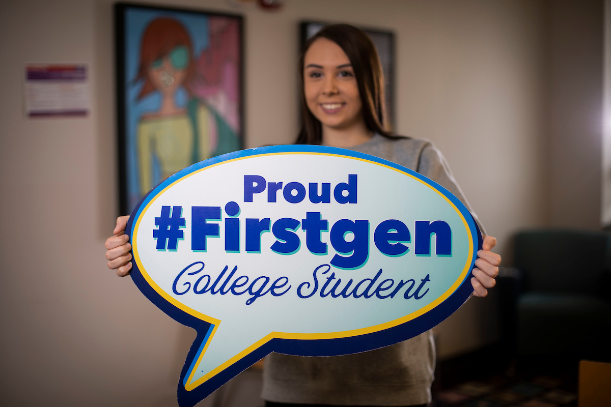 a photograph of a student holding a sign shaped like a talk bubble stating "proud #firstgen college student"