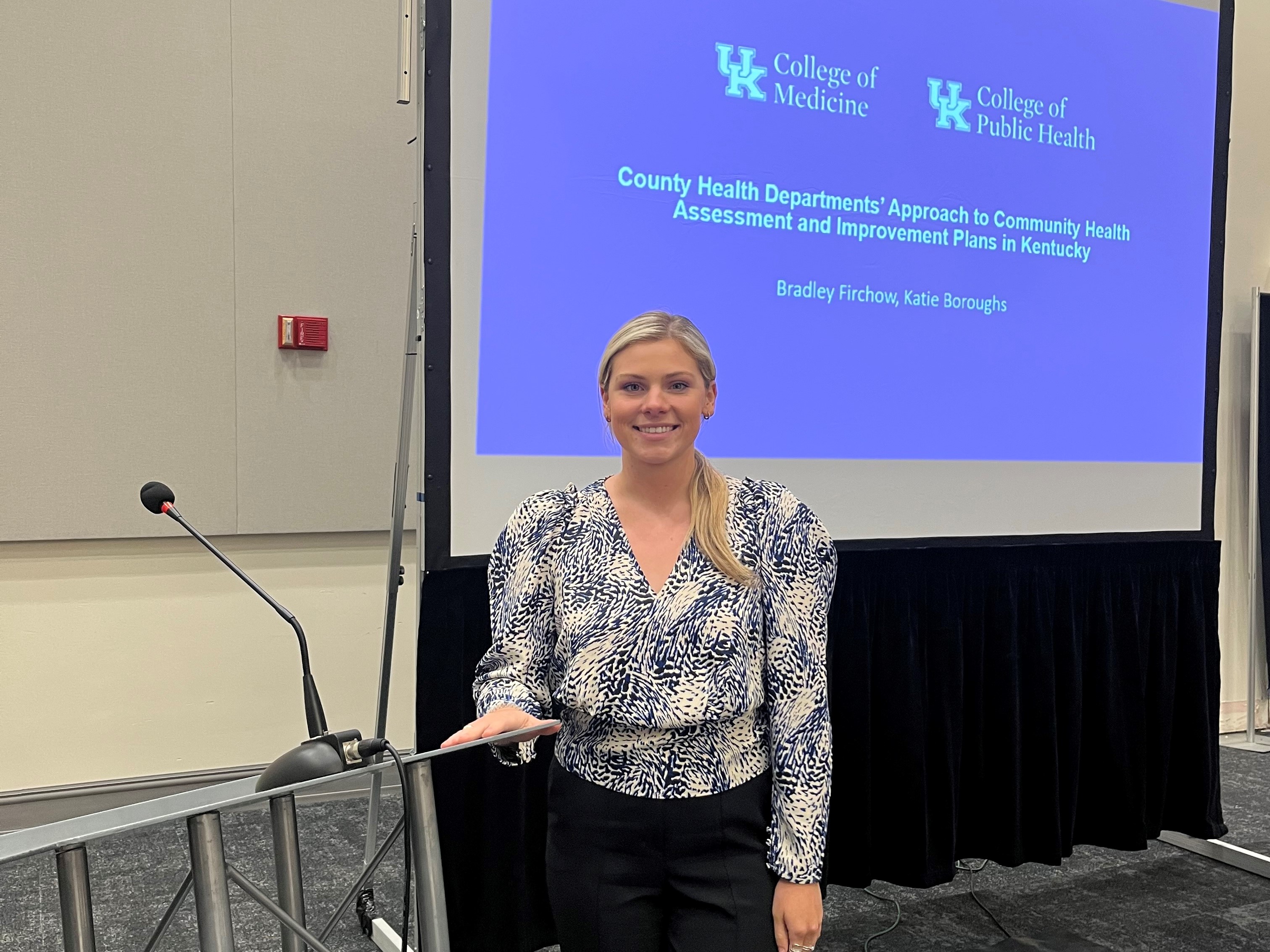 Pictured is Katie Boroughs standing in front of a podium and presentation backdrop