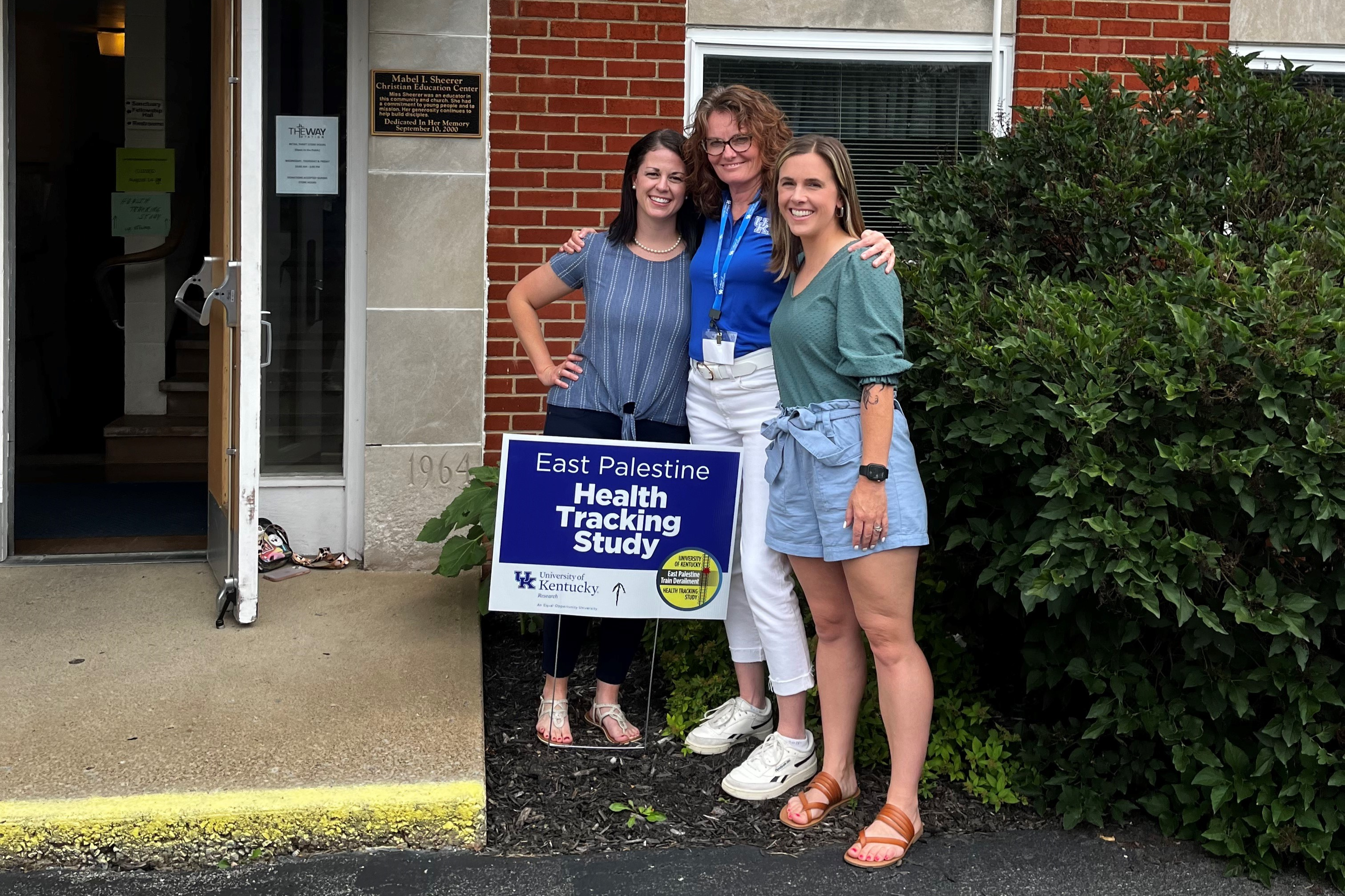 a photograph of Dr. Erin Haynes and two students in front a lawn sign that reads "East Palestine health tracking study"