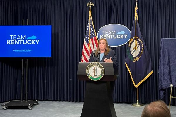 a photograph of Dana Quesinberry giving a speech behind a podium with a television projecting "Team Kentucky"