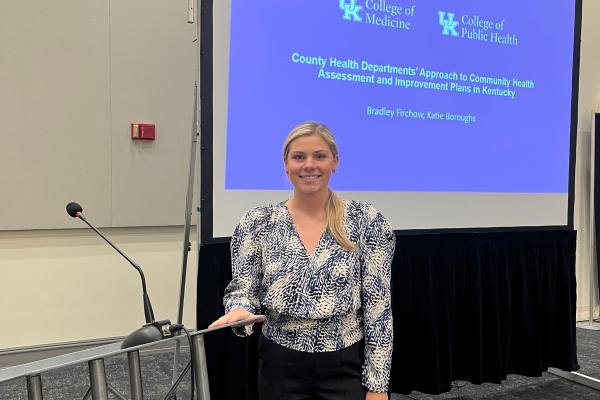 Pictured is Katie Boroughs standing in front of a podium and presentation backdrop