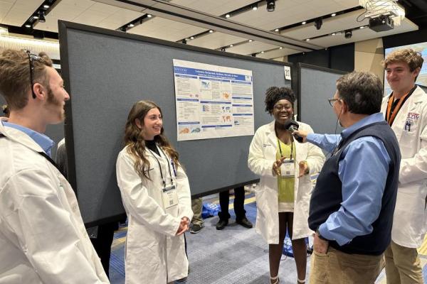 a photograph of students in white lab coats being interviewed in front of their poster