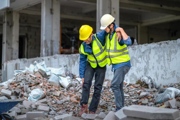 a photograph of two construction workers among rubble where one is helping the other walk from an injured leg