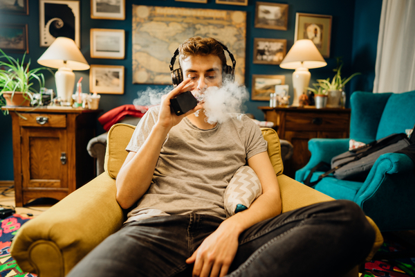 a stock photograph of a young man using a vape while listening on their headphones