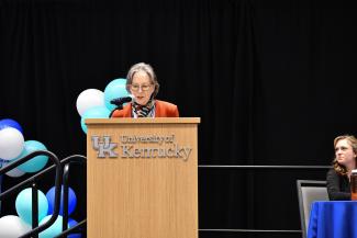 a photograph of Dr. Julia Costich speaking at a podium labeled “University of Kentucky”, on a stage of panelists