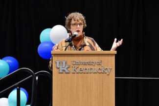 a photograph Dr. Connie White speaking at a podium labeled “University of Kentucky”, on a stage of panelists