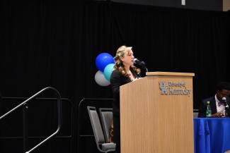 a photograph of Laura Eirich speaking at a podium labeled “University of Kentucky”, on a stage of panelists