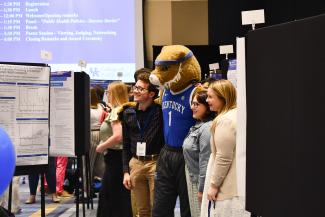 a photograph of three people smiling and posing with the Wildcat mascot, next to posters