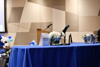 a photograph of Blue table with a tabletopper tent that reads “Public Health Showcase” next to a microphone and flower arrangement with white and light blue flowers