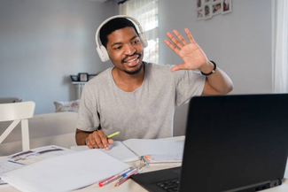 a stock photograph of a young person waving at their laptop computer while wearing over the ear headphones