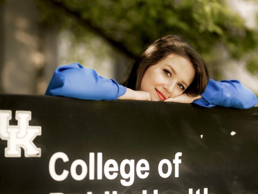 a photograph of Sarah Jane Robbins resting their head on a sign for the College of Public Health