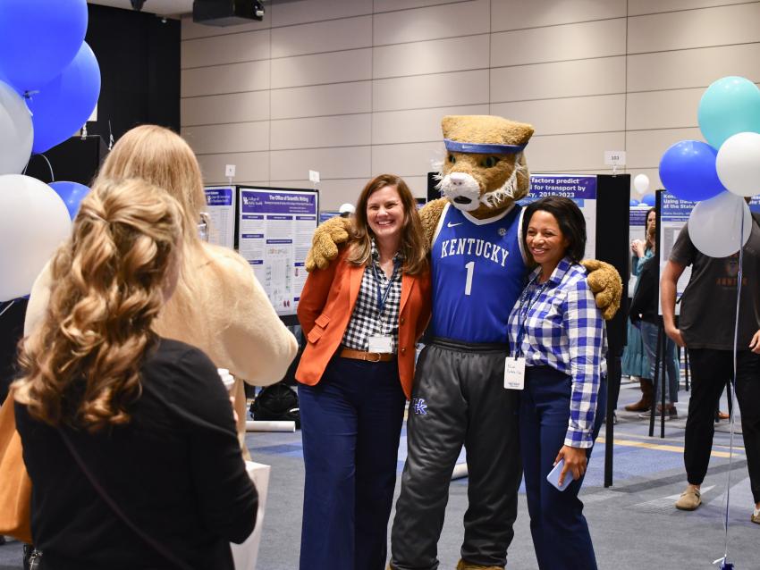 a photograph of a person taking a photo of two people posing with the Wildcat mascot. White, blue, and green balloons are on either side of the people.  