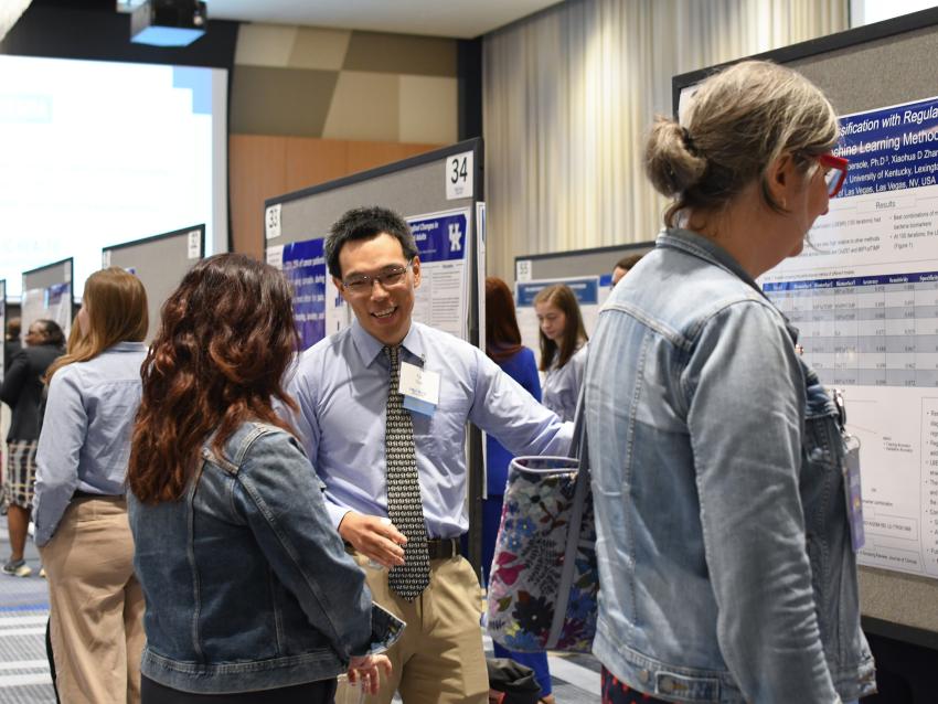 a photograph of a student talking about their research poster while pointing at it
