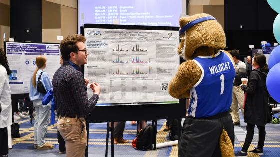 a photograph of a student at the Public Health Showcase showing the UK Wildcat mascot their presentation