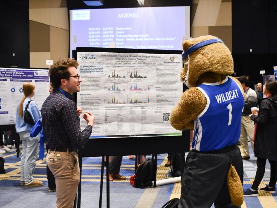 a photograph of a student at the Public Health Showcase showing the UK Wildcat mascot their presentation